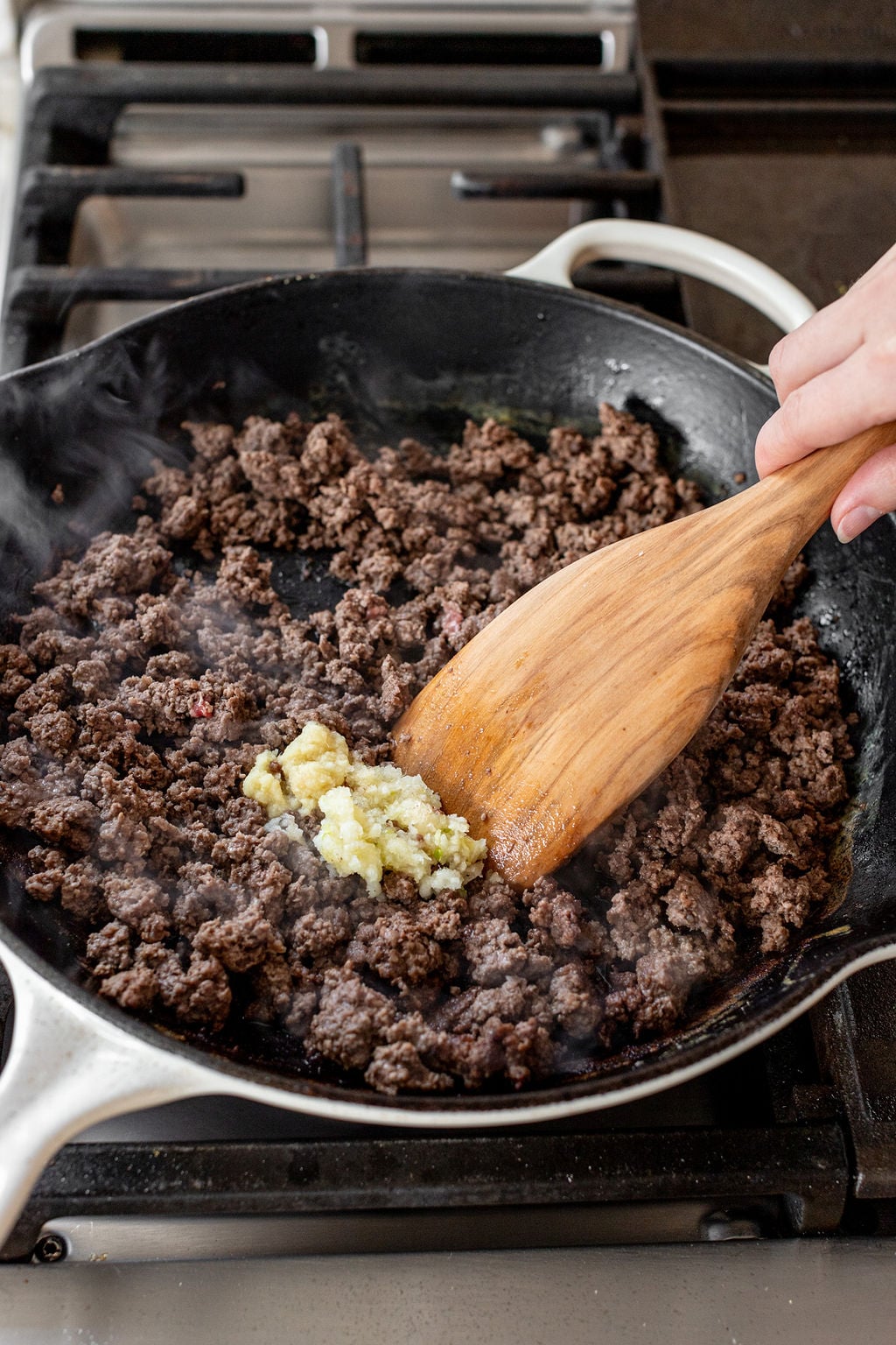 A large wooden spoon mixing garlic and ginger into a skillet of cooked ground beef.