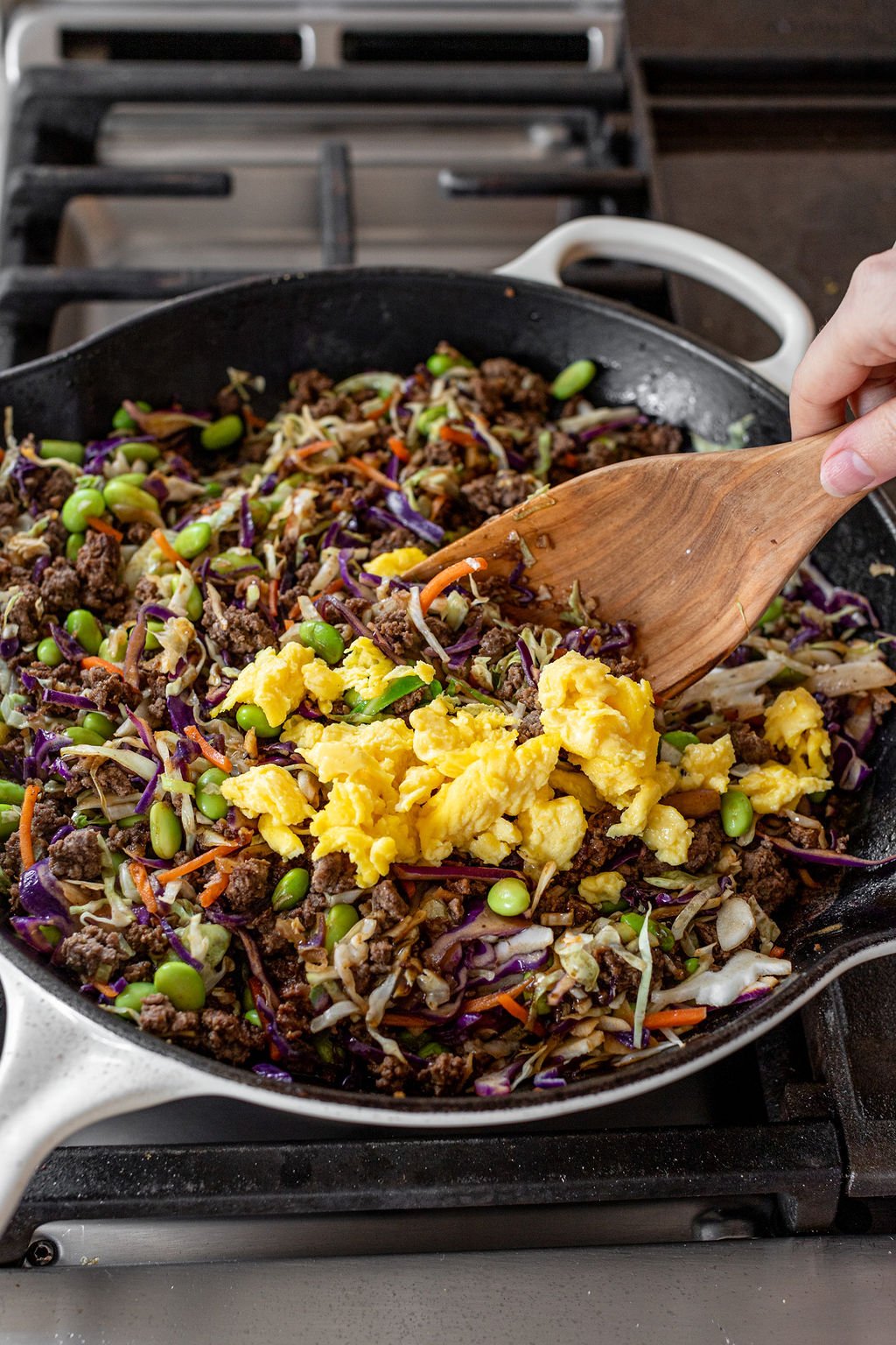 A hand mixing scrambled eggs into a large skillet with cooked ground beef, shredded cabbage and carrots, and edamame.