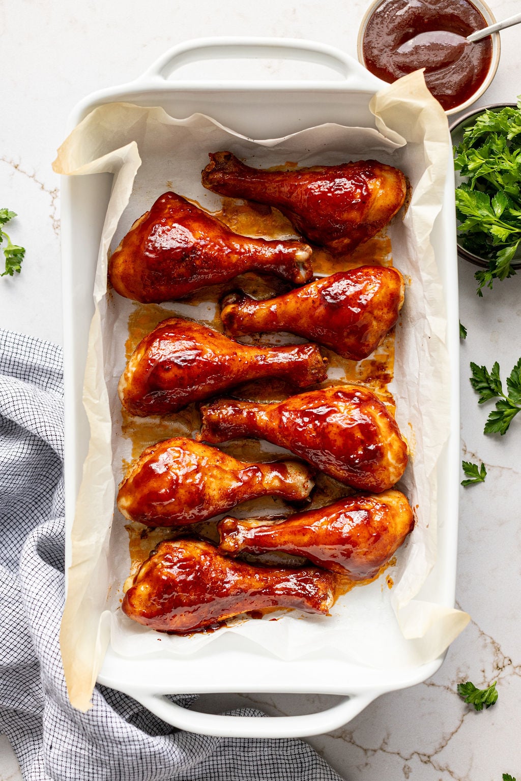 Eight drumsticks covered in glossy barbecue sauce in a dish next to a bowl of BBQ sauce.