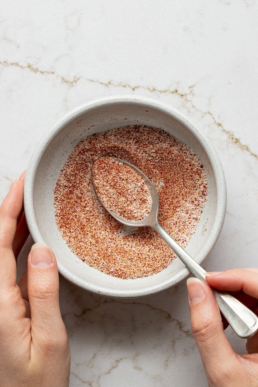 A hand mixing red and white spices in a bowl with a spoon.