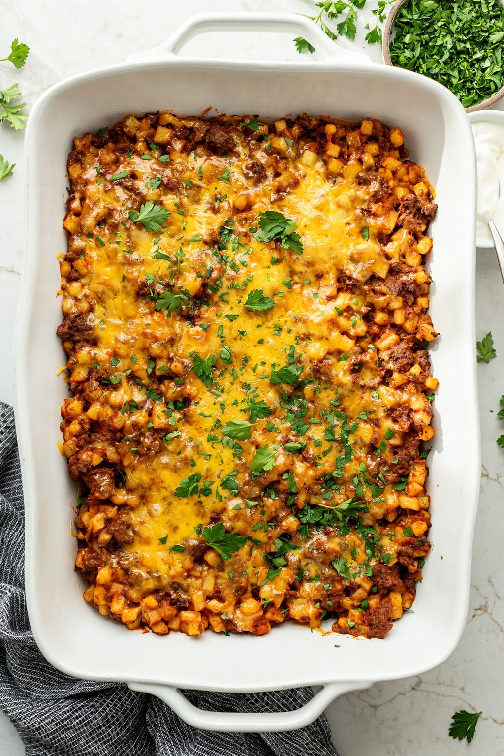 Chopped parsley spread on a baking dish filled with melted cheese, cooked ground beef, red sauce, and diced potatoes.