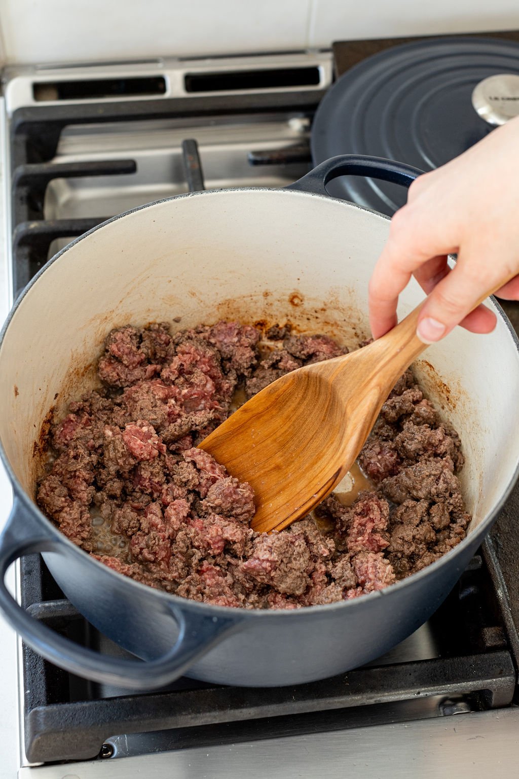 A hand stirring a pot of ground beef cooking in a large pot on a stove.