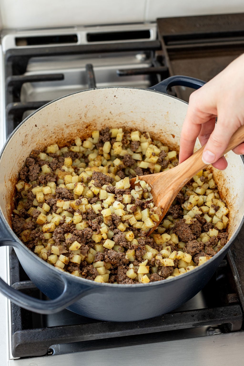 Cooked ground beef and small diced potatoes being stirred with a large spoon in a metal pot.
