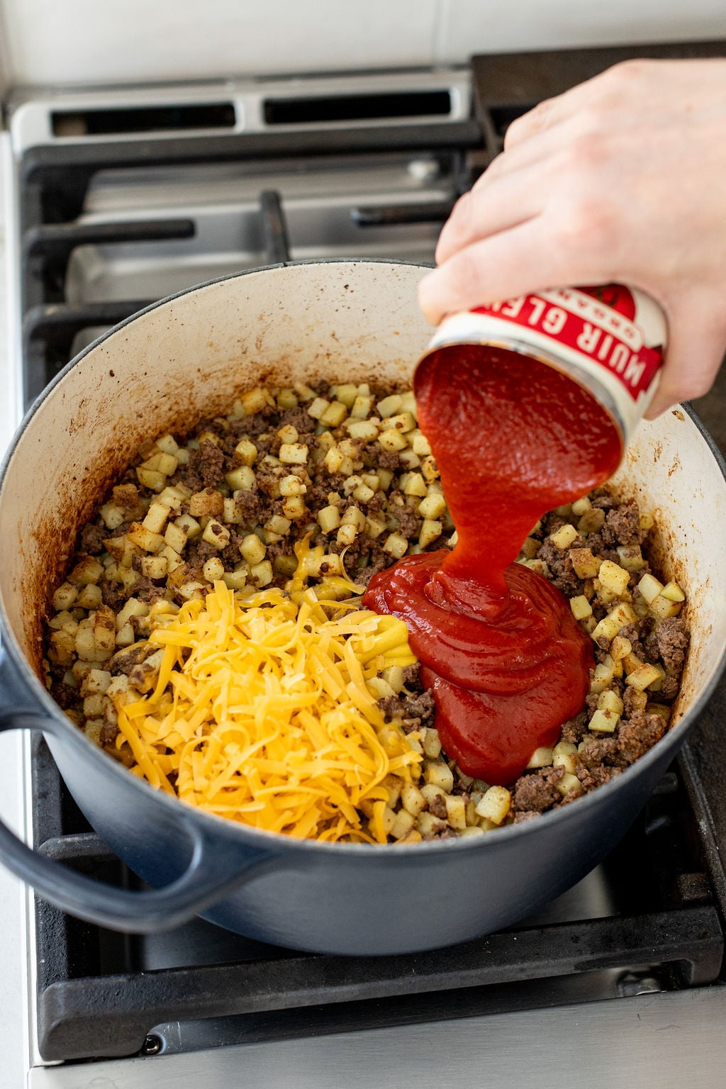 A hand pouring tomato sauce into a pot with ground beef, potatoes, and shredded cheese.