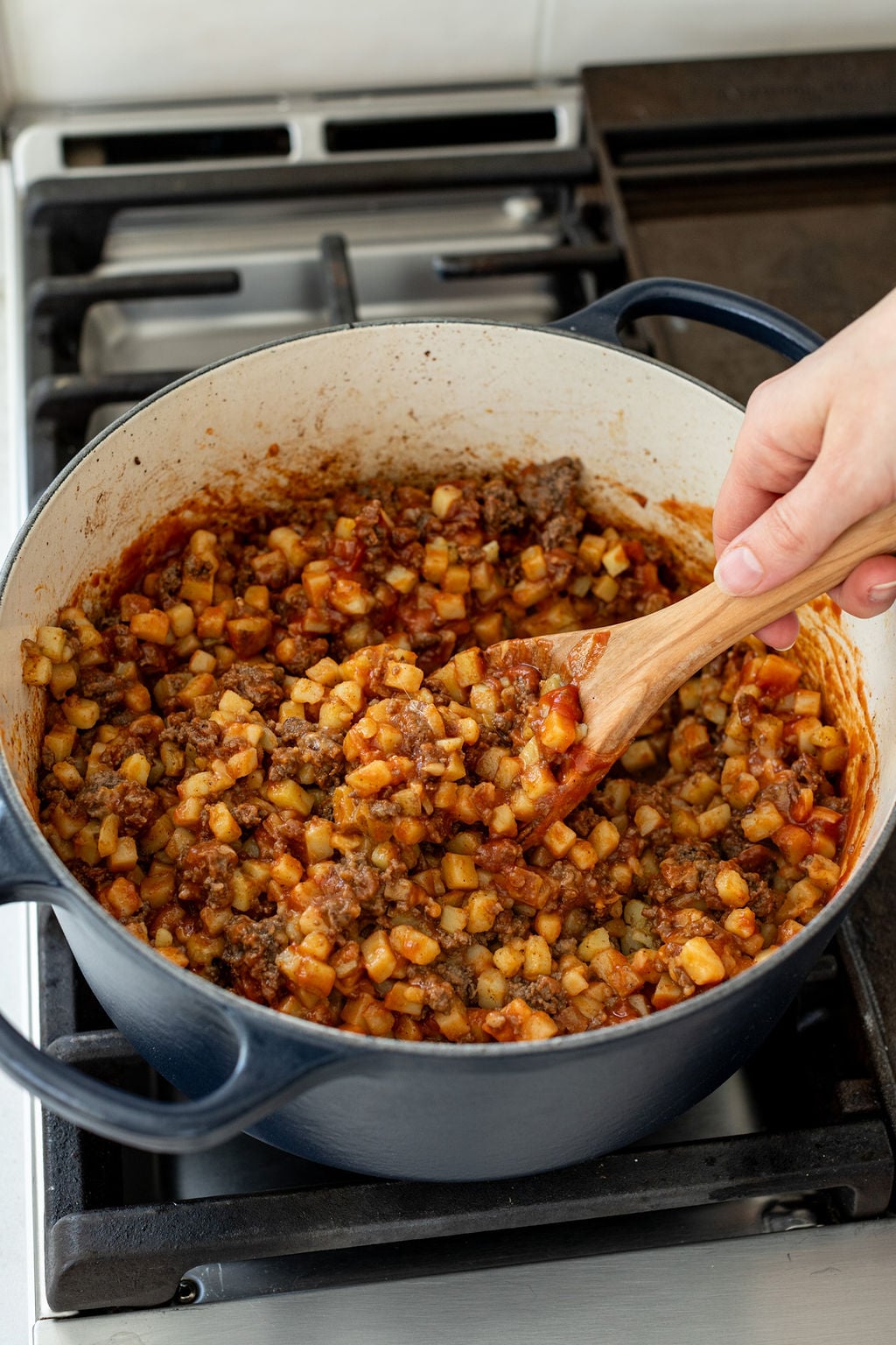 A hand stirring cooked diced potatoes, ground beef, and red tomato sauce with a large wooden spoon in a pot.