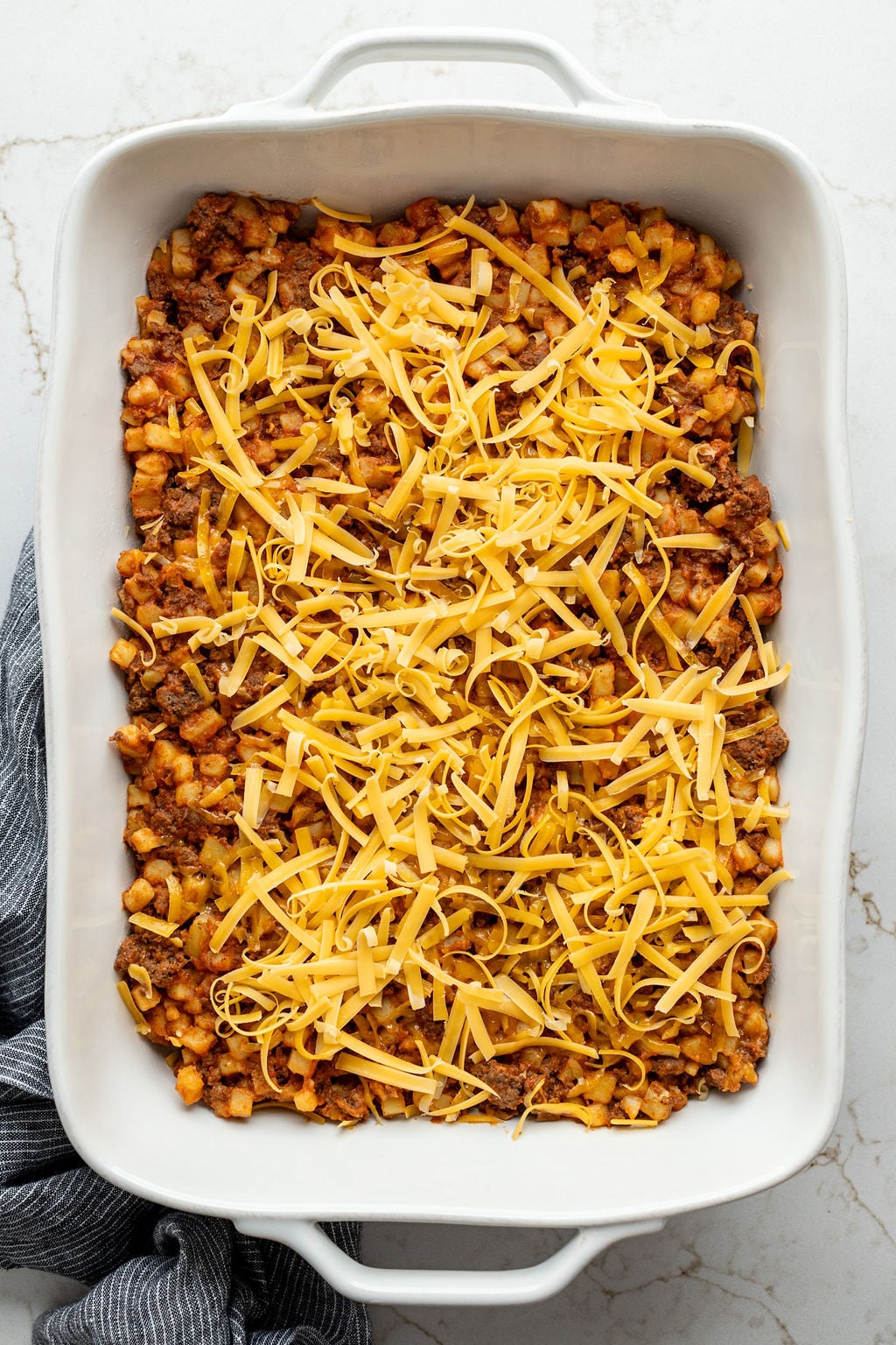 Cooked ground beef and diced potatoes spread in a white baking dish and topped with shredded cheese.