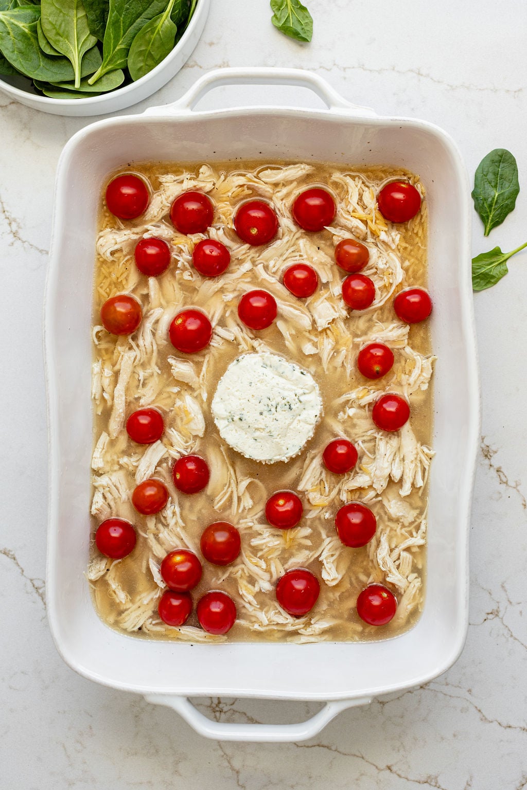 Cherry tomatoes and a block of Boursin cheese sitting on top of stock, shredded chicken, and orzo in a baking dish.