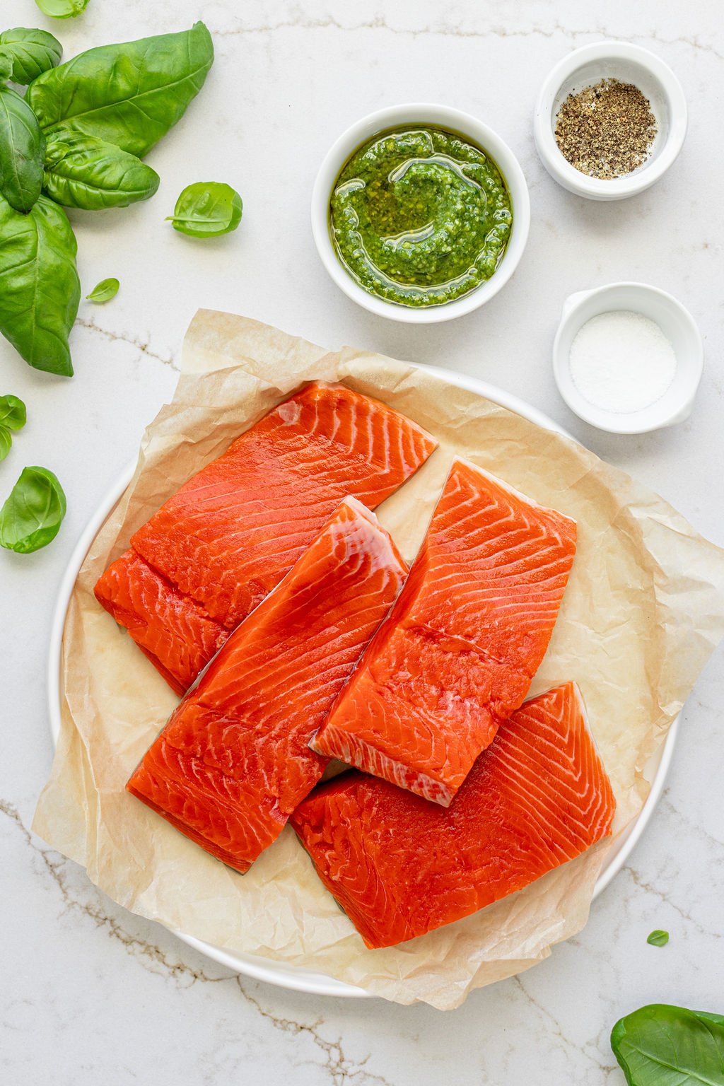 Four salmon filets on a plate next to bowls of salt, pepper, and green pesto.