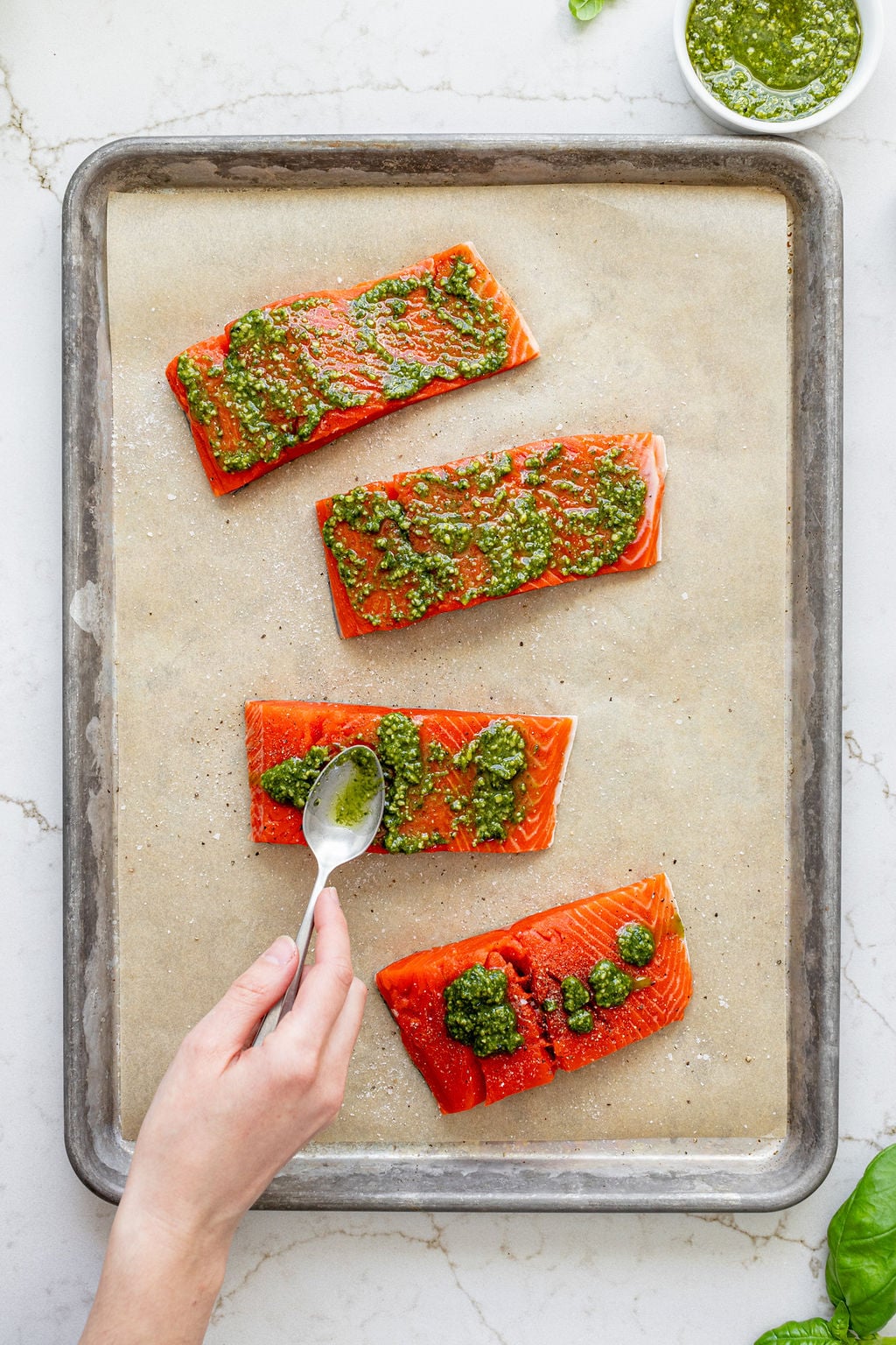 A hand spreading a creamy green spread onto salmon filets with a spoon.