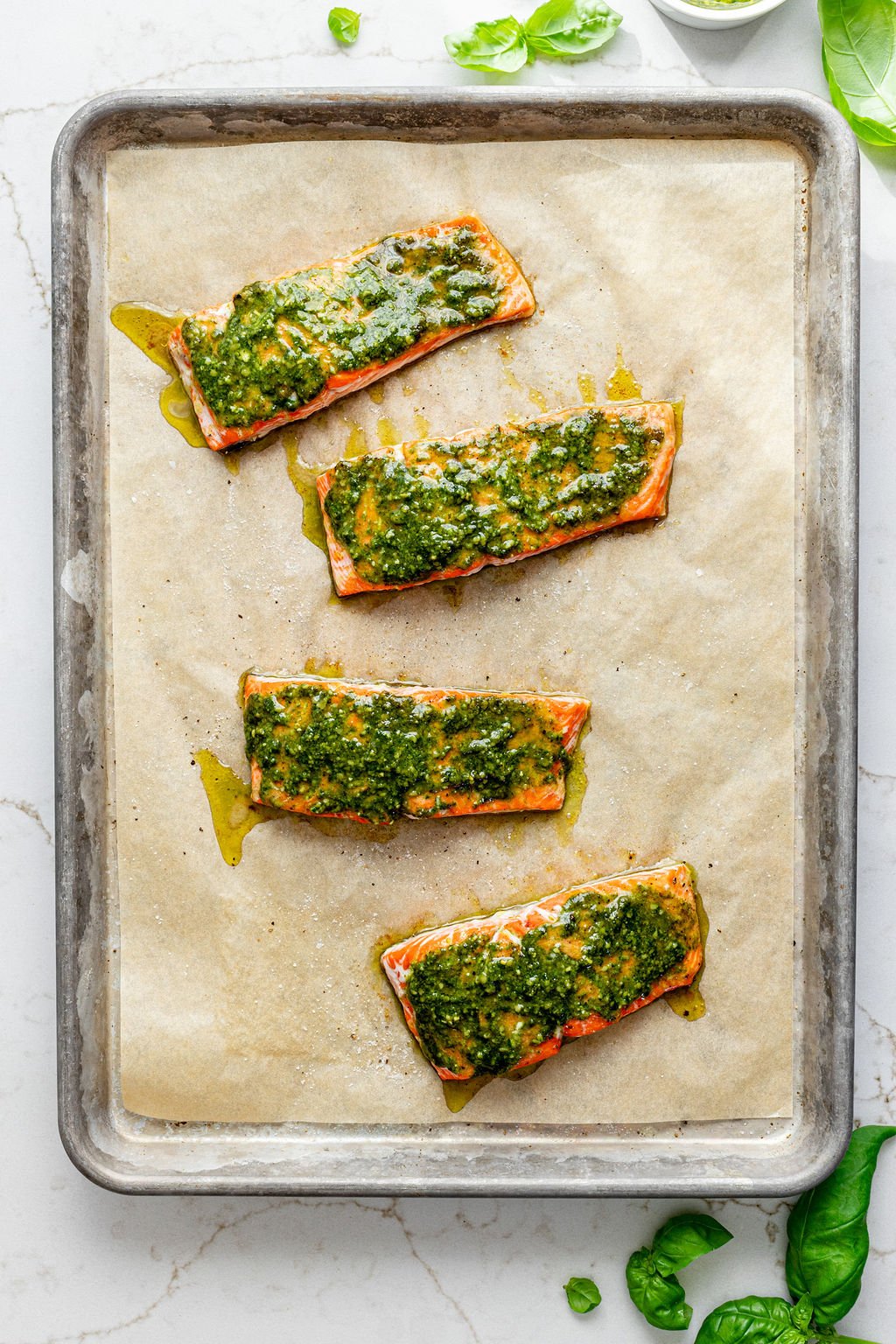 Four baked filets of salmon on a metal baking sheet topped with creamy pesto.