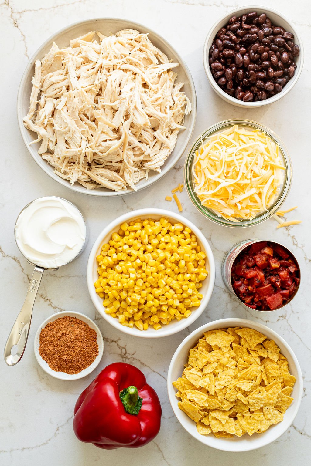 Bowls of cooked shredded chicken, diced tomatoes, black beans, sour cream, shredded cheese, corn chips, and corn on a table.