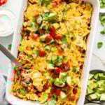 A baking dish filled with a melty and creamy taco casserole, topped with crushed corn chips and avocado.