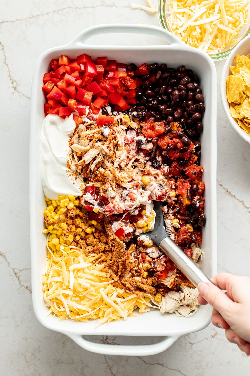 A hand mixing cooked shredded chicken, diced tomatoes, black beans, sour cream, shredded cheese, and corn in a baking dish.