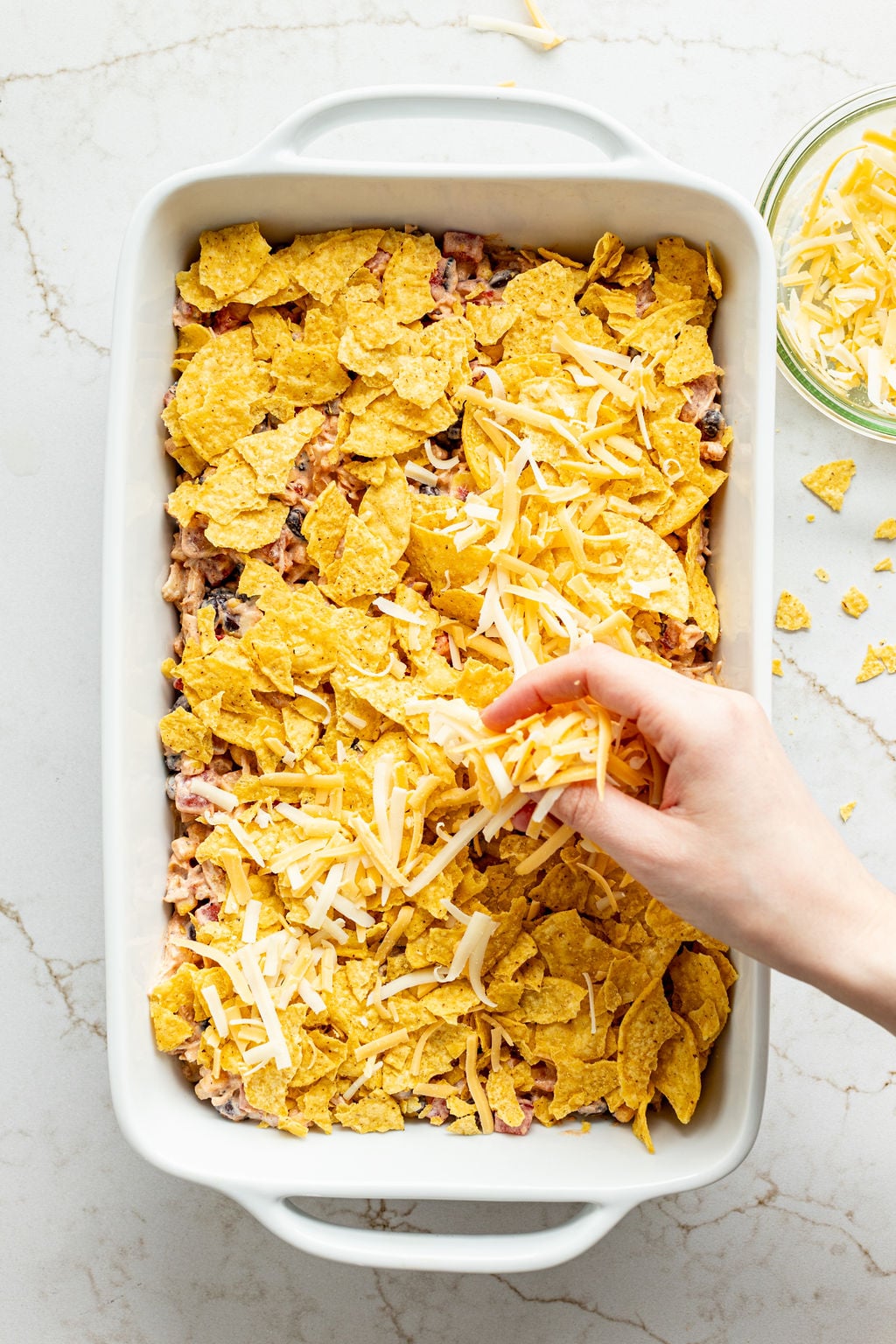 A hand sprinkling shredded cheese into a casserole dish.