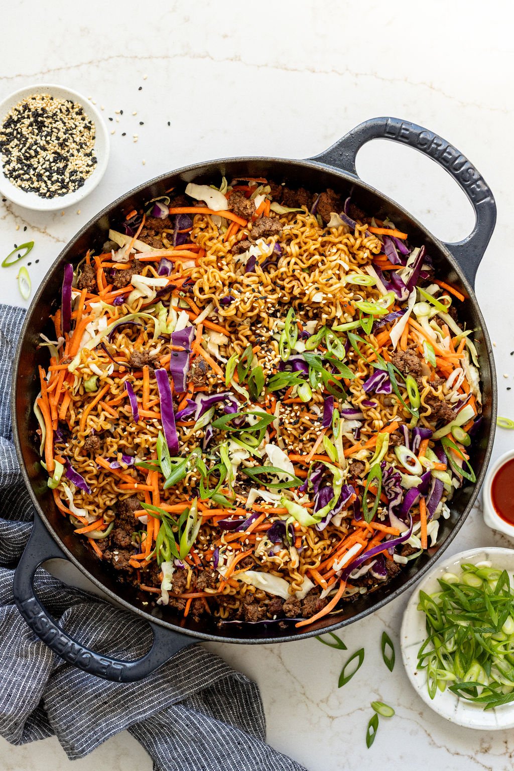 A large cast iron skillet on a table filled with creamy ramen, brown sauce, ground beef, and shredded vegetables.