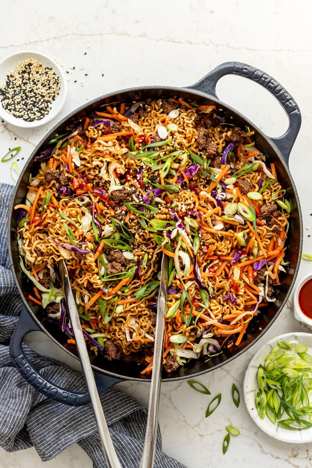 Metal tongs sitting in a skillet of ramen, shredded vegetables, coleslaw mix, and cooked beef crumbles.
