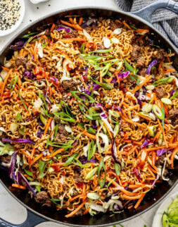 Bowls of sesame seeds and green onions next to a skillet filled with cooked ramen, brown sauce, ground beef, and slaw mix,