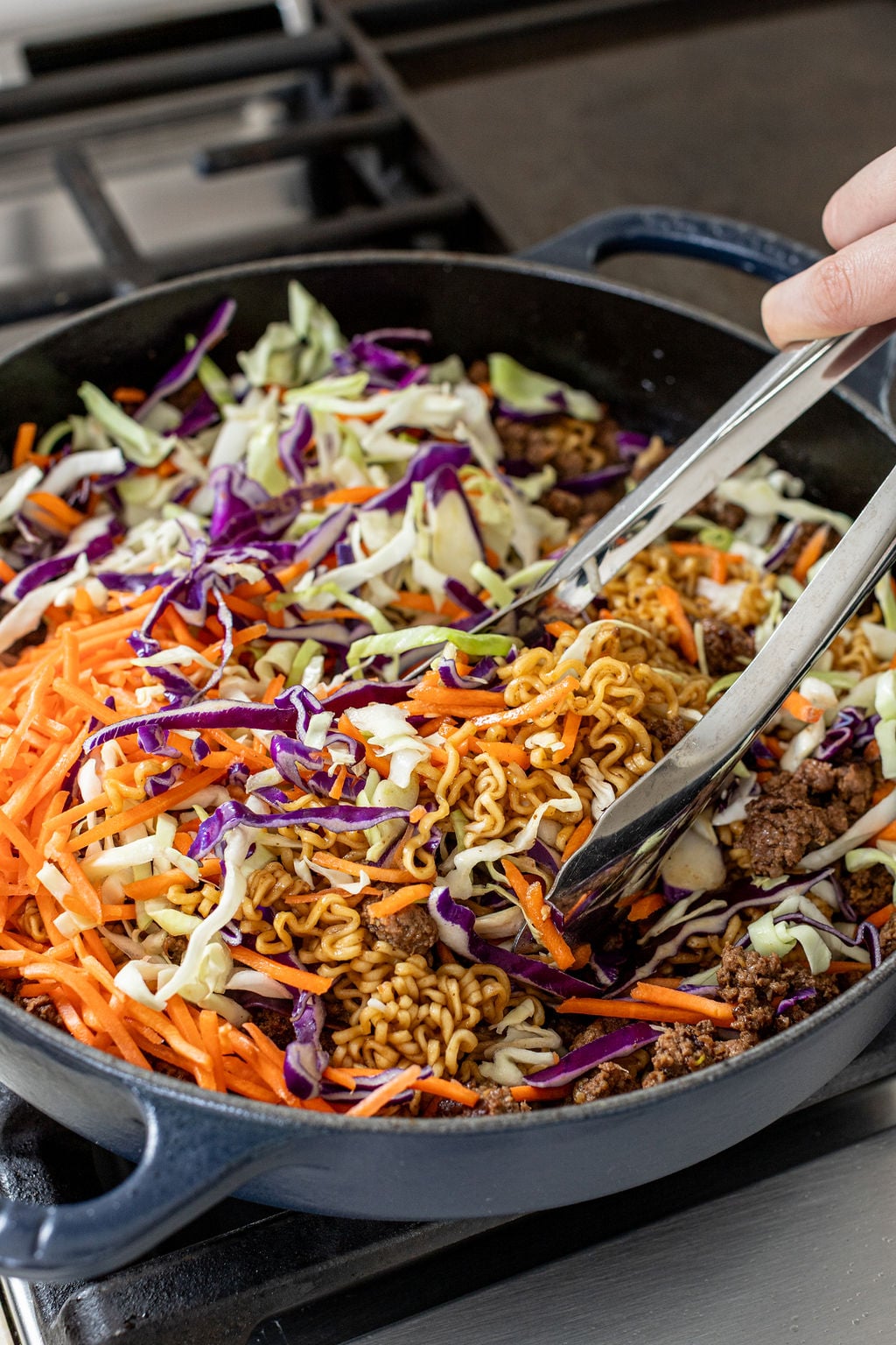 Hands mixing ramen noodles with coleslaw mix in a skillet with metal tongs.