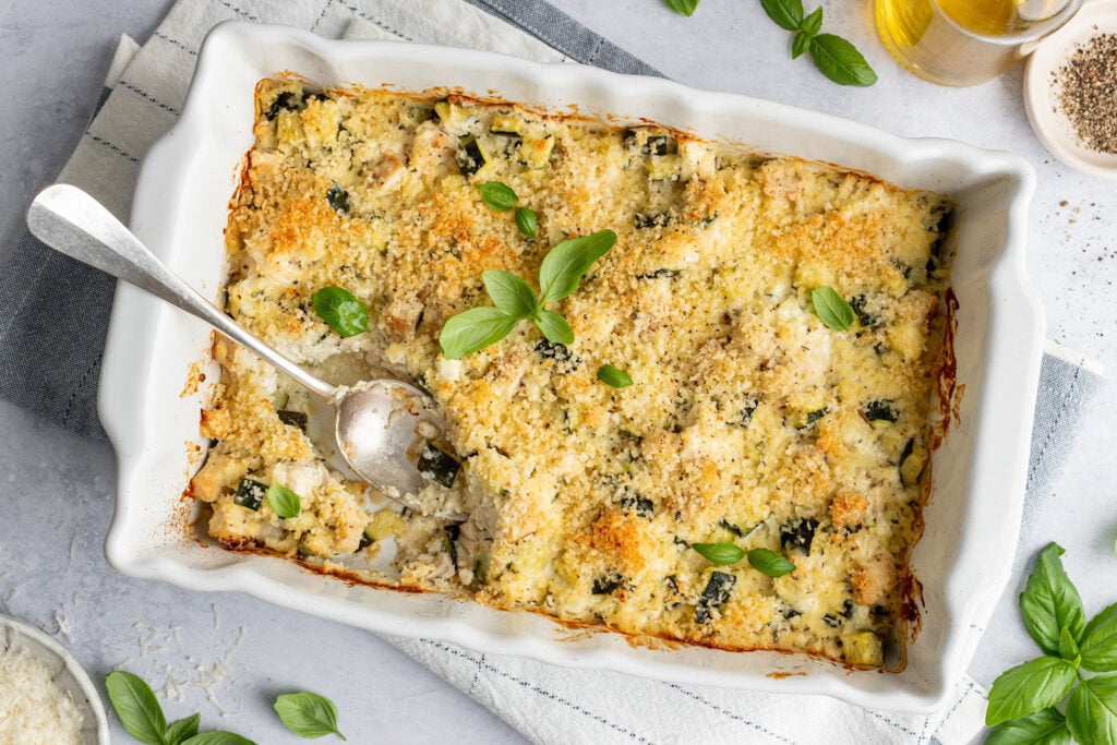 White baking dish on top of a towel with a spoon and a baked zucchini casserole covered in breadcrumbs and topped with basil. Baking dish is surrounded basil leaves, small bowls of pepper and shredded cheese, and a jar of oil.