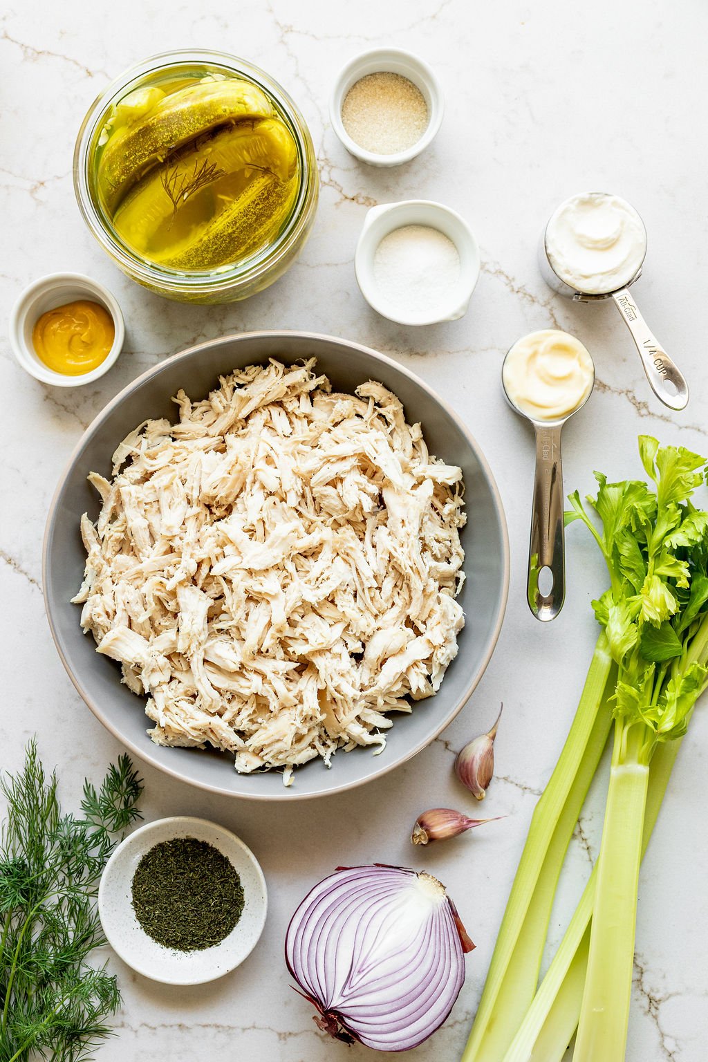 Bowls of shredded chicken, pickles, spices, mayo, and mustard on a table.