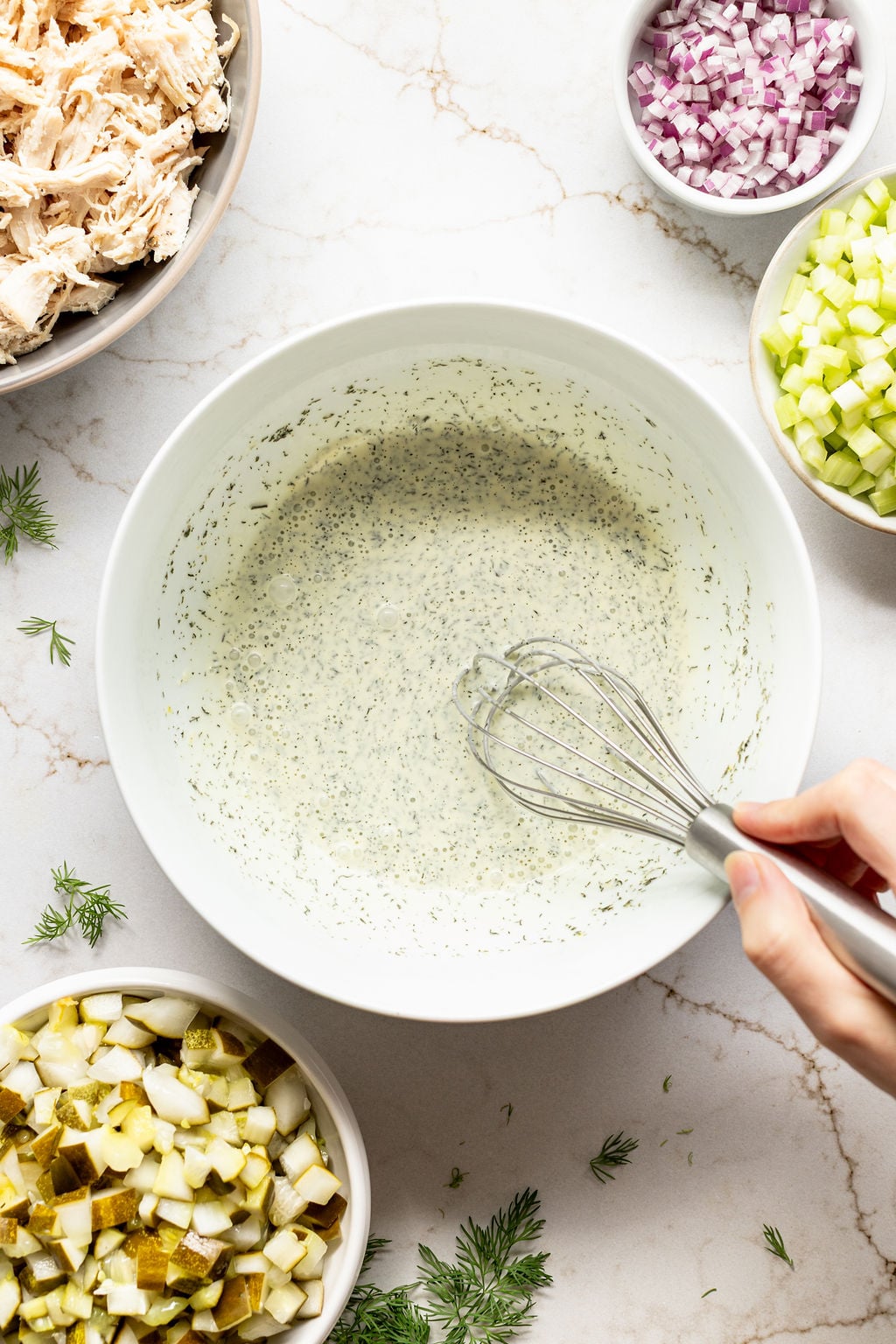 A hand whisking mustard, yogurt, mayonnaise, spices, sugar, dill, and pickle juice in a bowl.