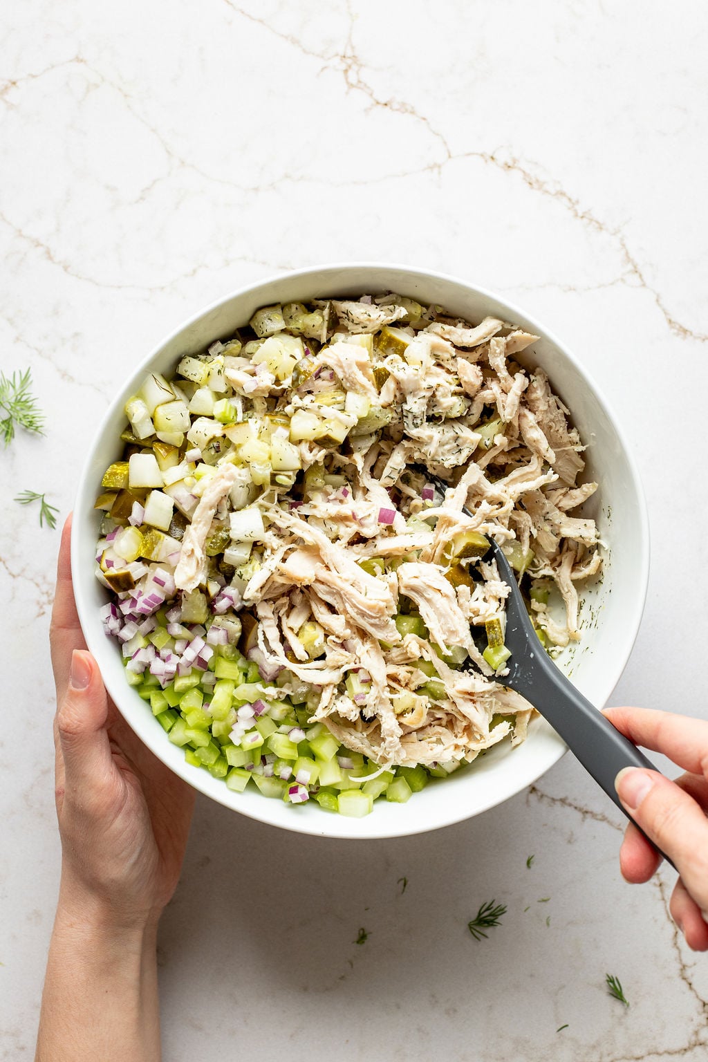 A hand stirring diced pickles, red onion, celery, and shredded chicken in a bowl.