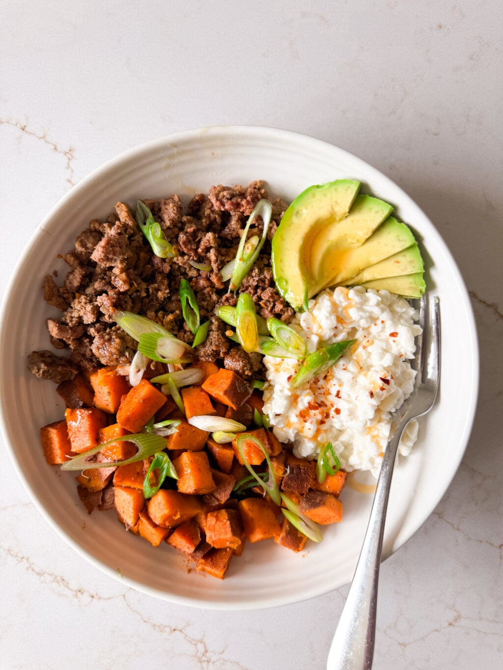 A bowl with cooked ground beef, diced sweet potatoes, cottage cheese, and sliced avocado.