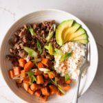 A bowl with cooked ground beef, diced sweet potatoes, cottage cheese, and sliced avocado.