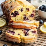 Two stacked slices of glazed blueberry cake on a cooling rack next to the rest of the loaf cake.