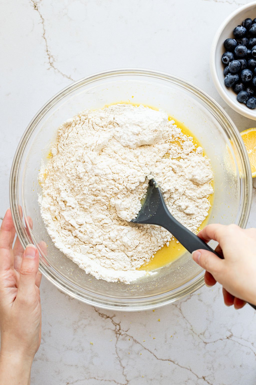 A hand stirring flour into a wet yellow mixture in a mixing bowl.