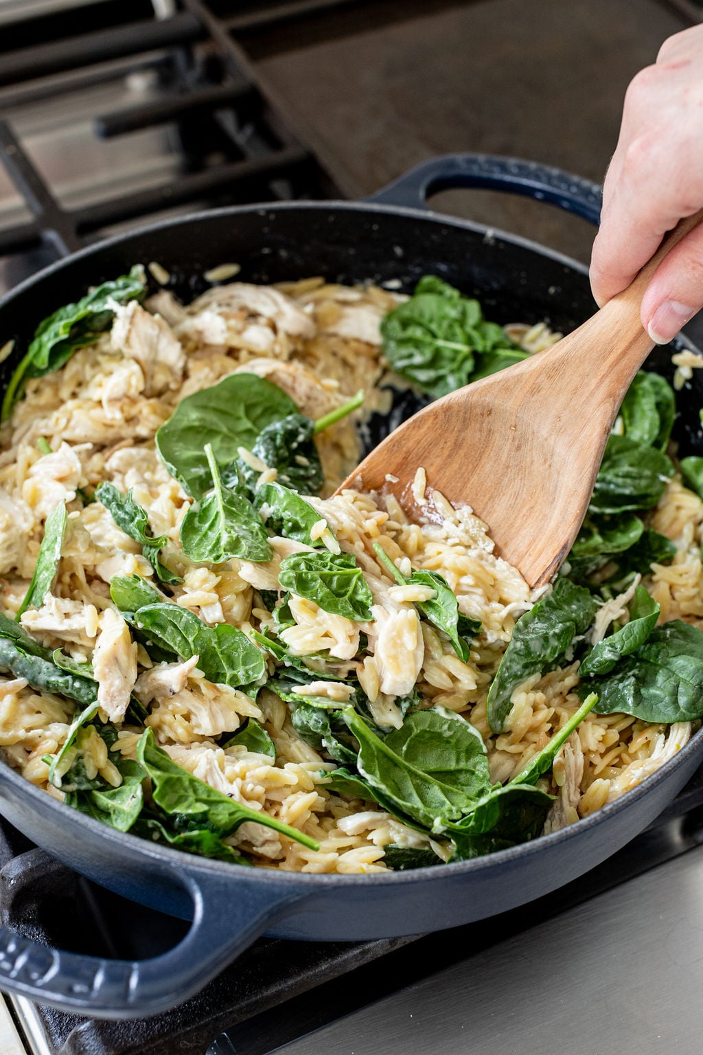 A hand stirring spinach into creamy orzo and chicken with a wooden spoon.