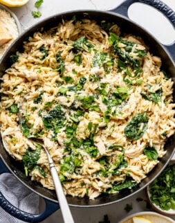 A metal spoon sitting in a large skillet filled with creamy orzo, shredded chicken, spinach, cream cheese, and parmesan.