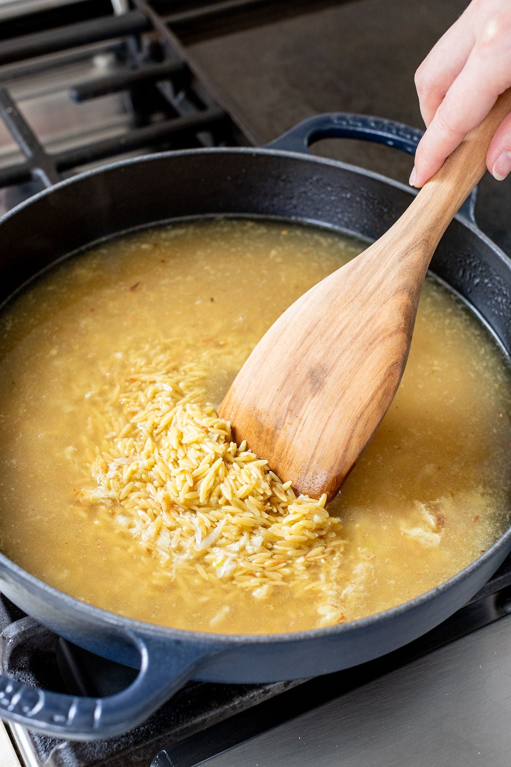 A wooden spoon stirring broth and orzo in a large skillet.