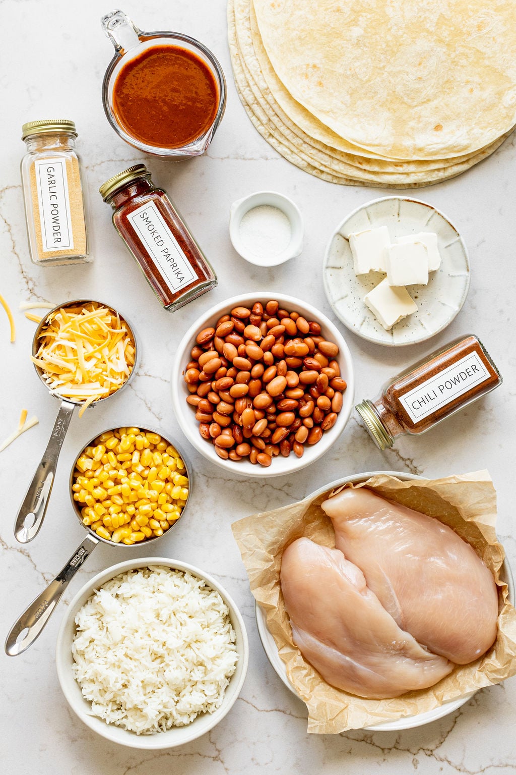 Bowls of beans, chicken breast, corn, shredded cheese, spices, and rice next to tortillas.