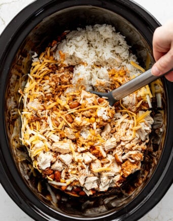A hand stirring shredded cheese, cooked chicken, rice, enchilada sauce, beans, and corn in a crockpot.