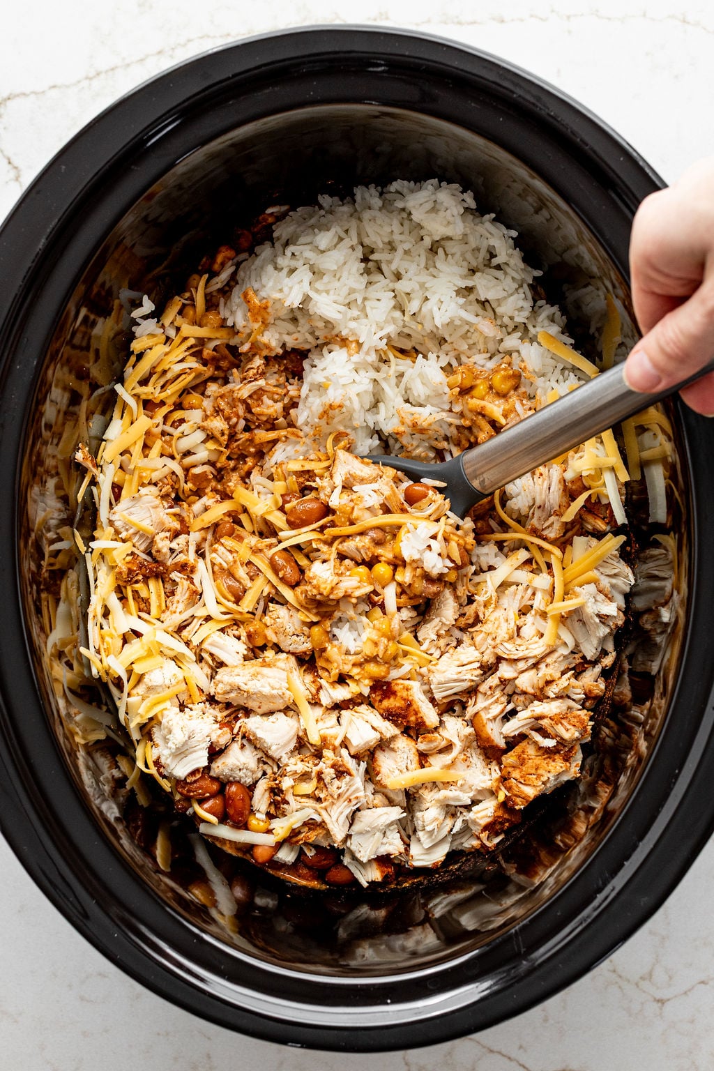 A hand stirring shredded cheese, cooked chicken, rice, enchilada sauce, beans, and corn in a crockpot.