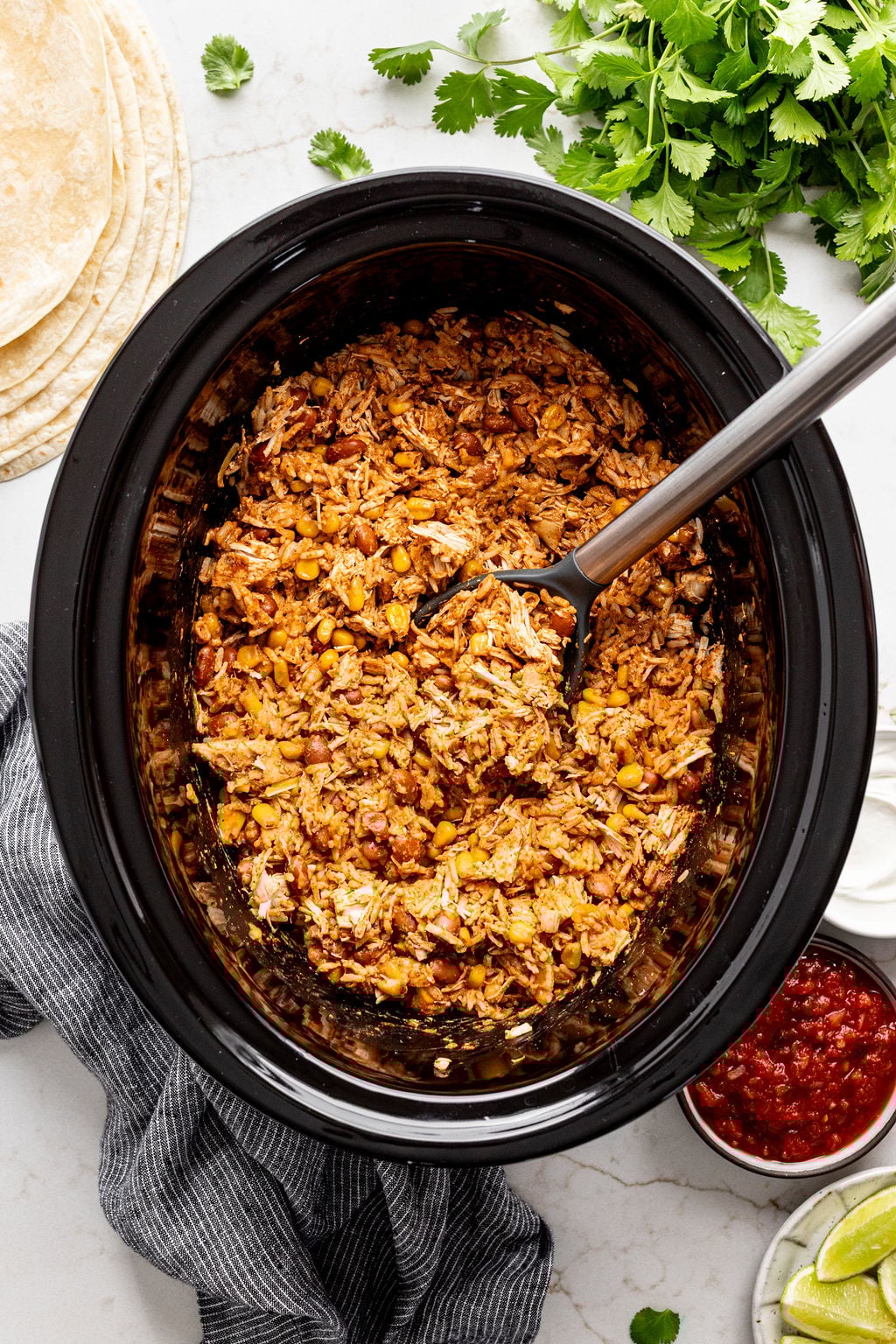 Tortillas and bowls of salsa and sour cream next to a crockpot filled with A spoon in a slow cooker filled with spiced shredded chicken, enchilada sauce, pinto beans, corn, and taco seasoning.