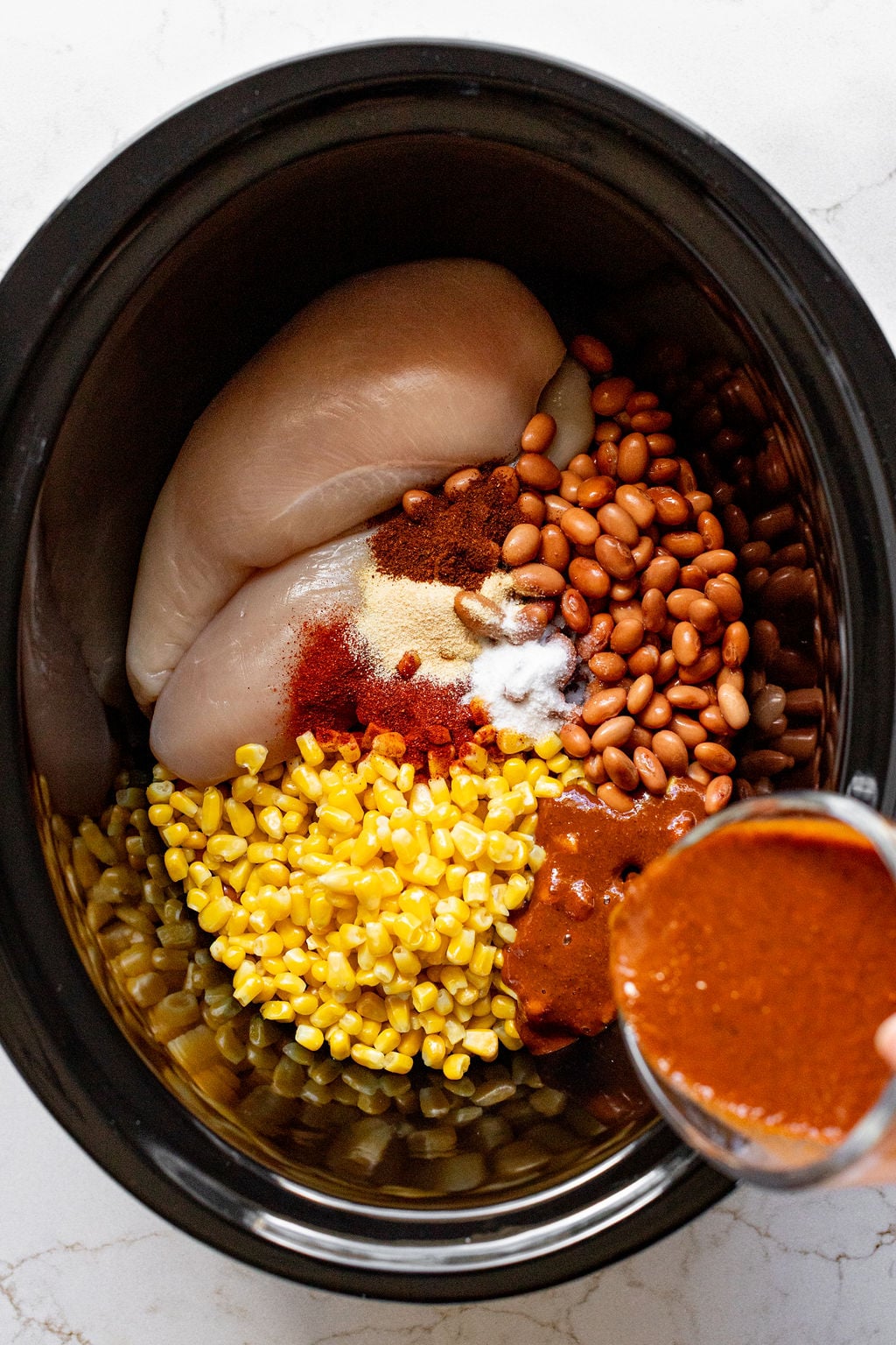 A slow cooker filled with chicken breasts, corn, pinto beans, and seasonings.