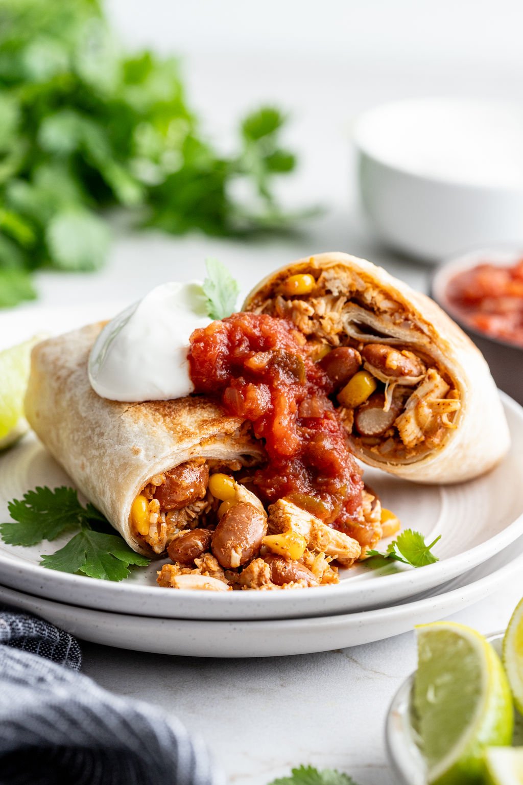 Two halves of a chicken burrito stacked on a plate topped with sour cream and salsa.