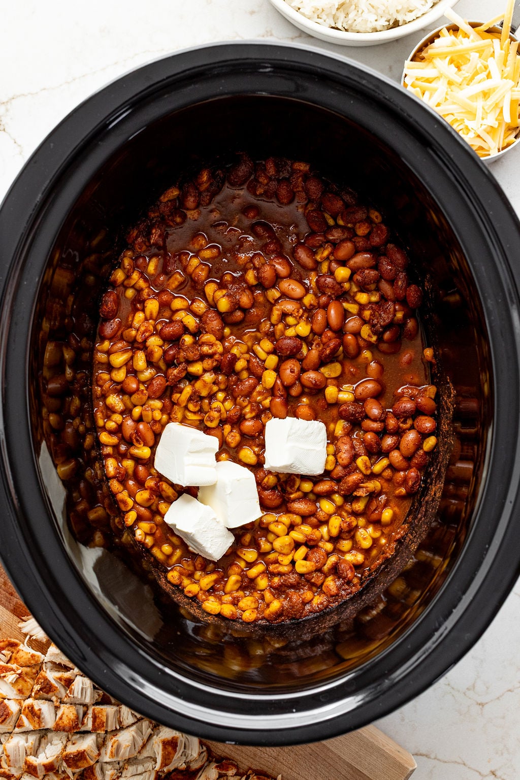 Small blocks of cream cheese on top of saucy beans and corn in a slow cooker.