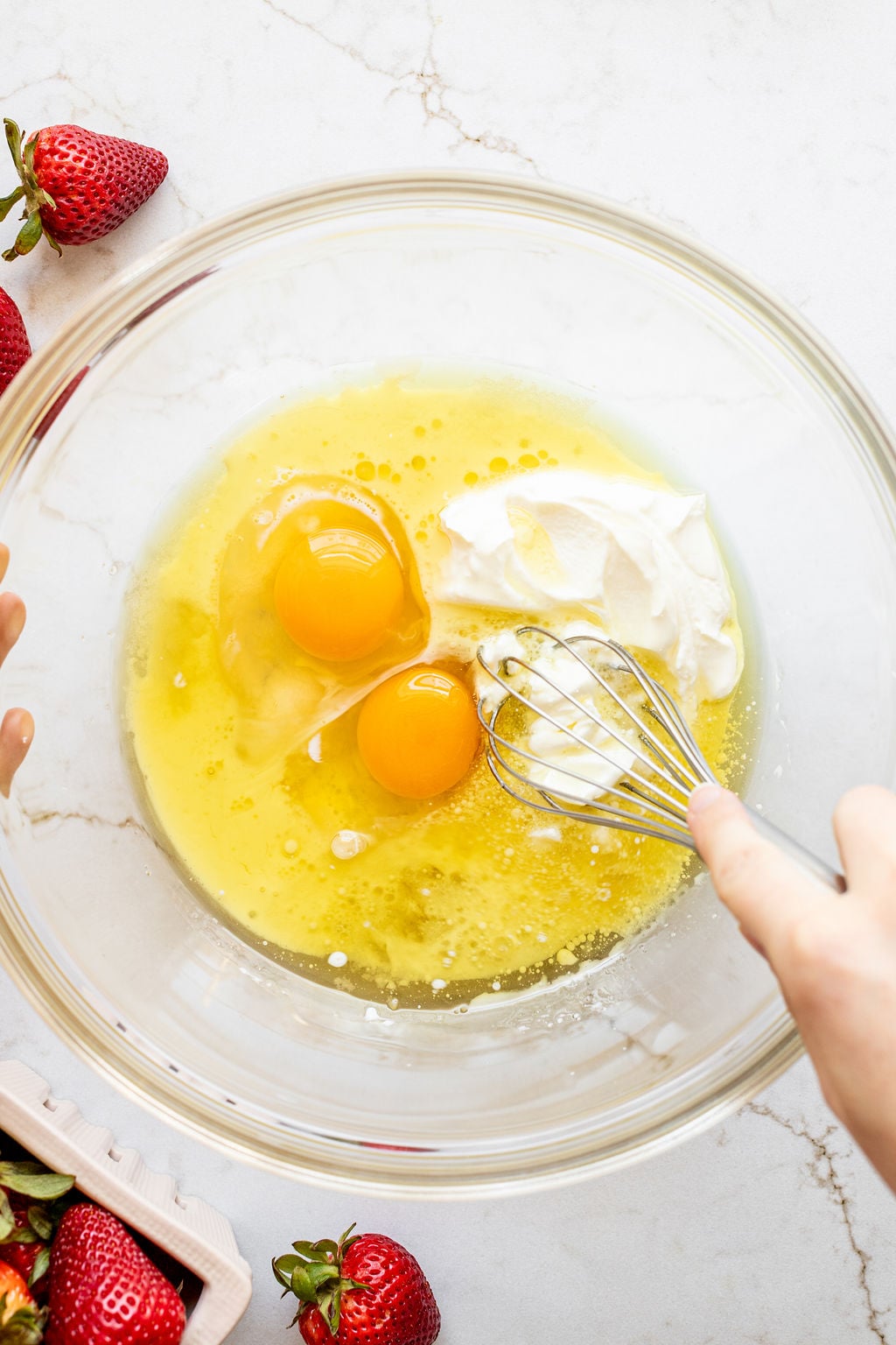 A hand whisking a bowl of oil, eggs, milk, and Greek yogurt,