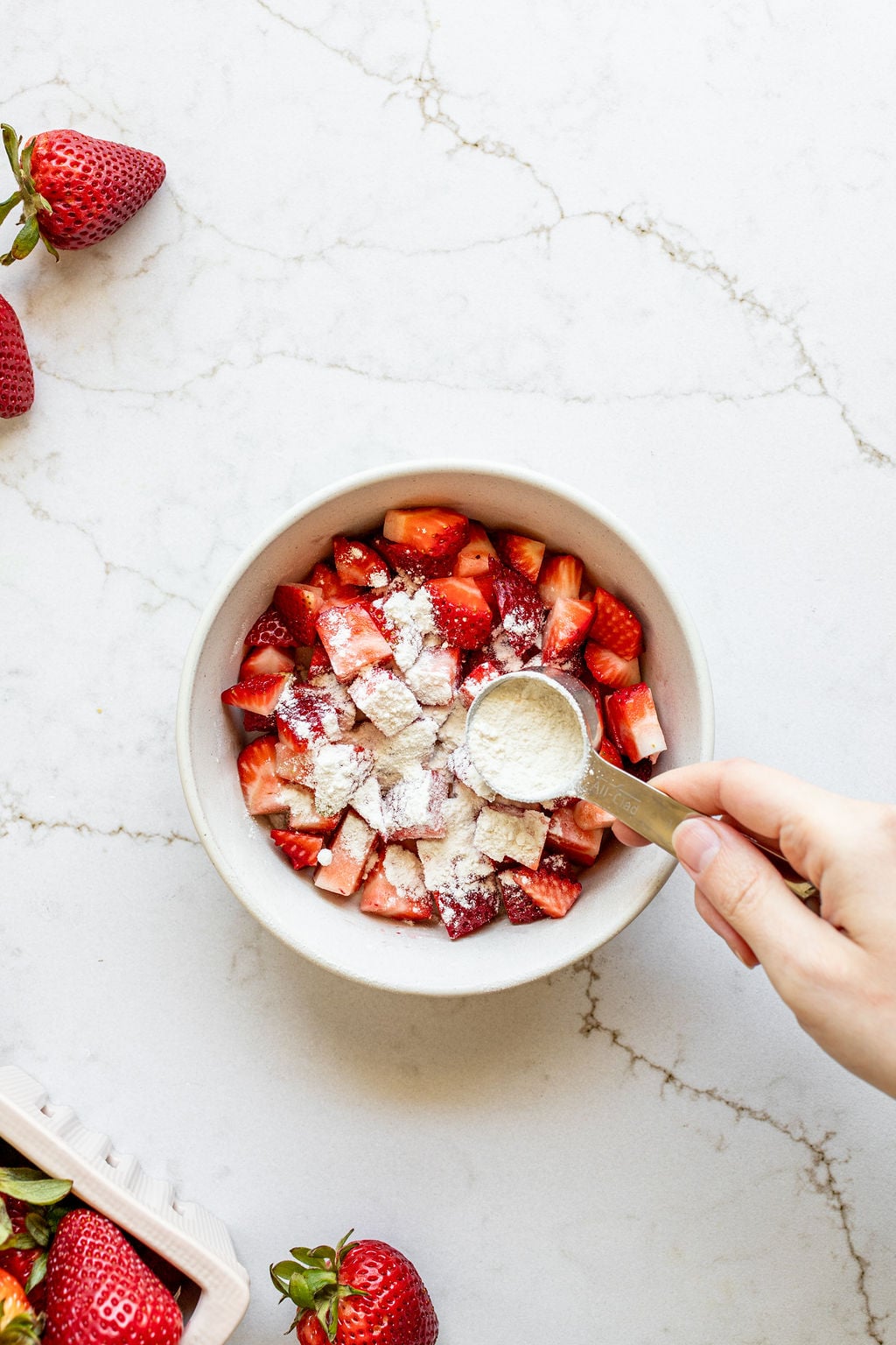 A hand pouring flour onto chopped strawberries in a bowl.