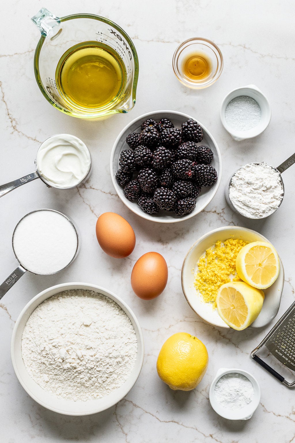 Bowls of blackberries, flour, sugar, Greek yogurt, oil, vanilla extract, and lemon zest next to eggs and more lemons.