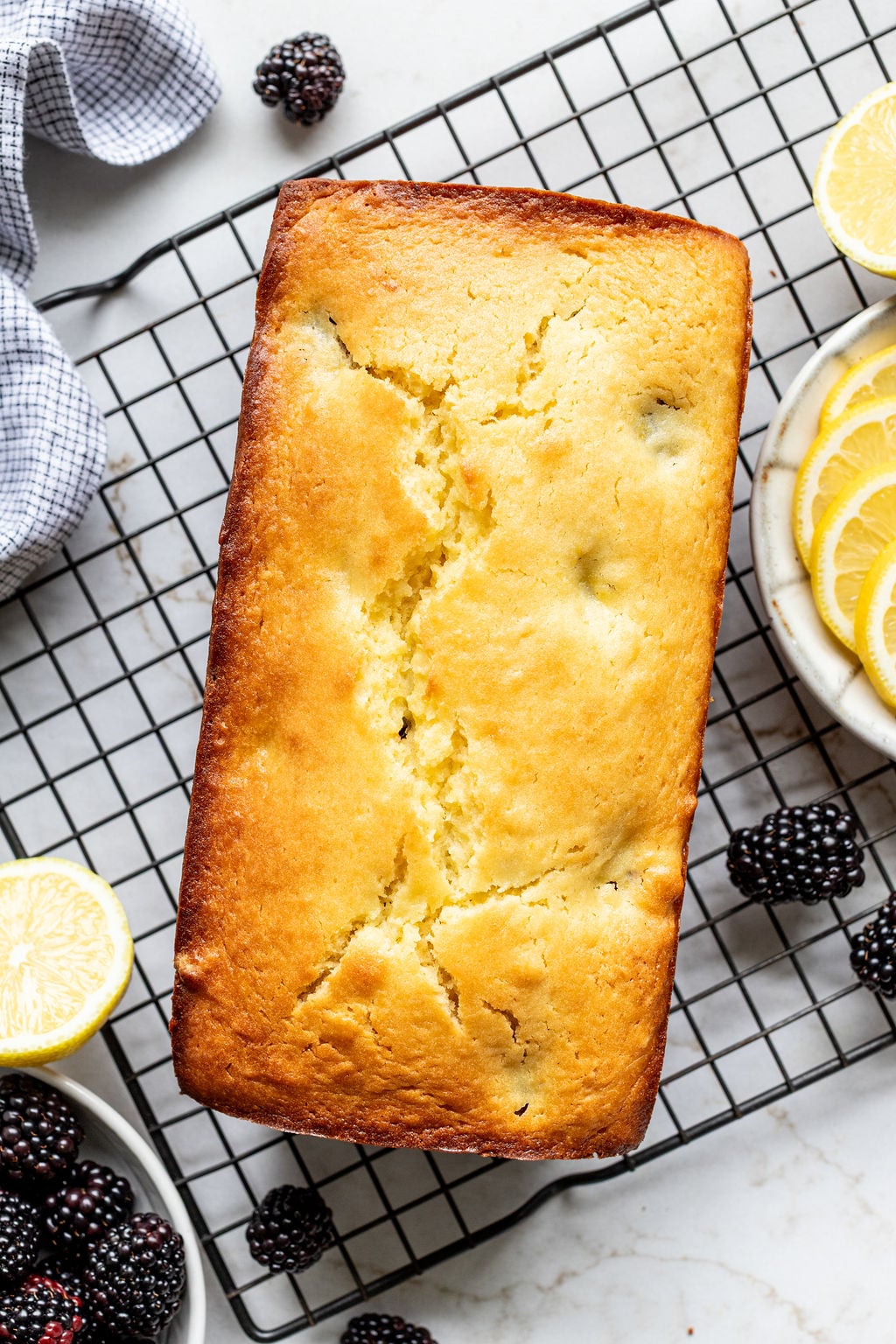 A baked loaf cake on a cooling rack next to blackberries and lemon slices.