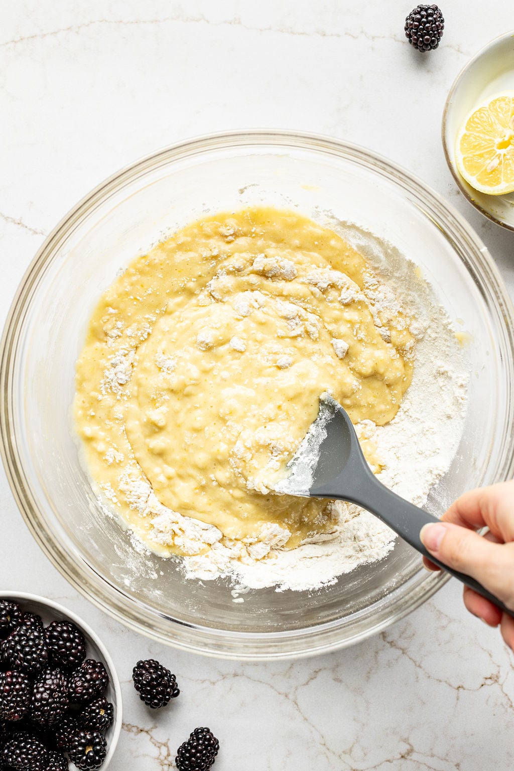 A hand stirring flour into yellow cake batter in a mixing bowl.