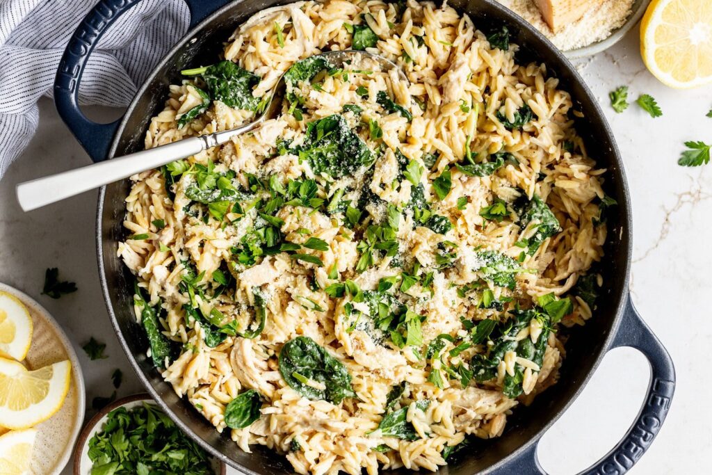 Lemon slices and a block of parmesan next to a skillet filled with creamy orzo, shredded chicken, spinach, and cream cheese.
