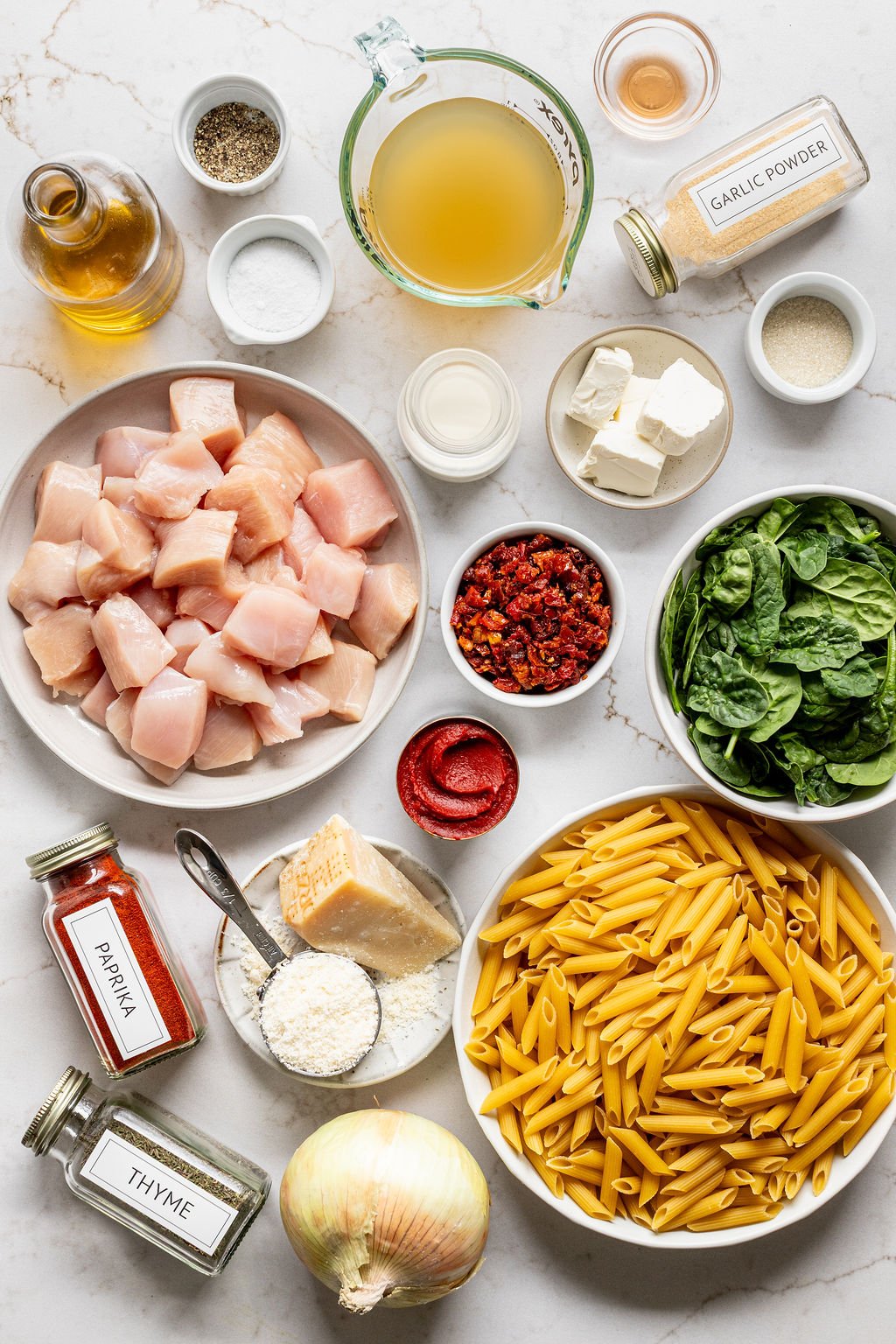 Bowls of cubed chicken, pasta, baby spinach, chicken broth, sun dried tomatoes, tomato pasta, creamy cheese, parmesan, and spices on a table.