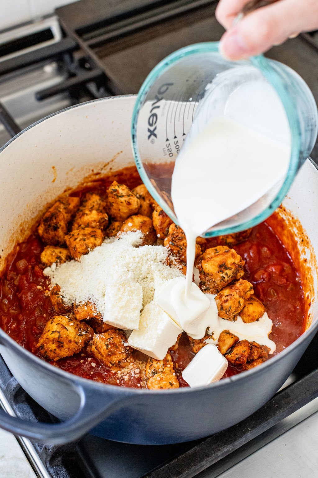 A hand pouring heavy cream into a pot with chunky chicken, red sauce, cubed cream cheese, and parmesan cheese.