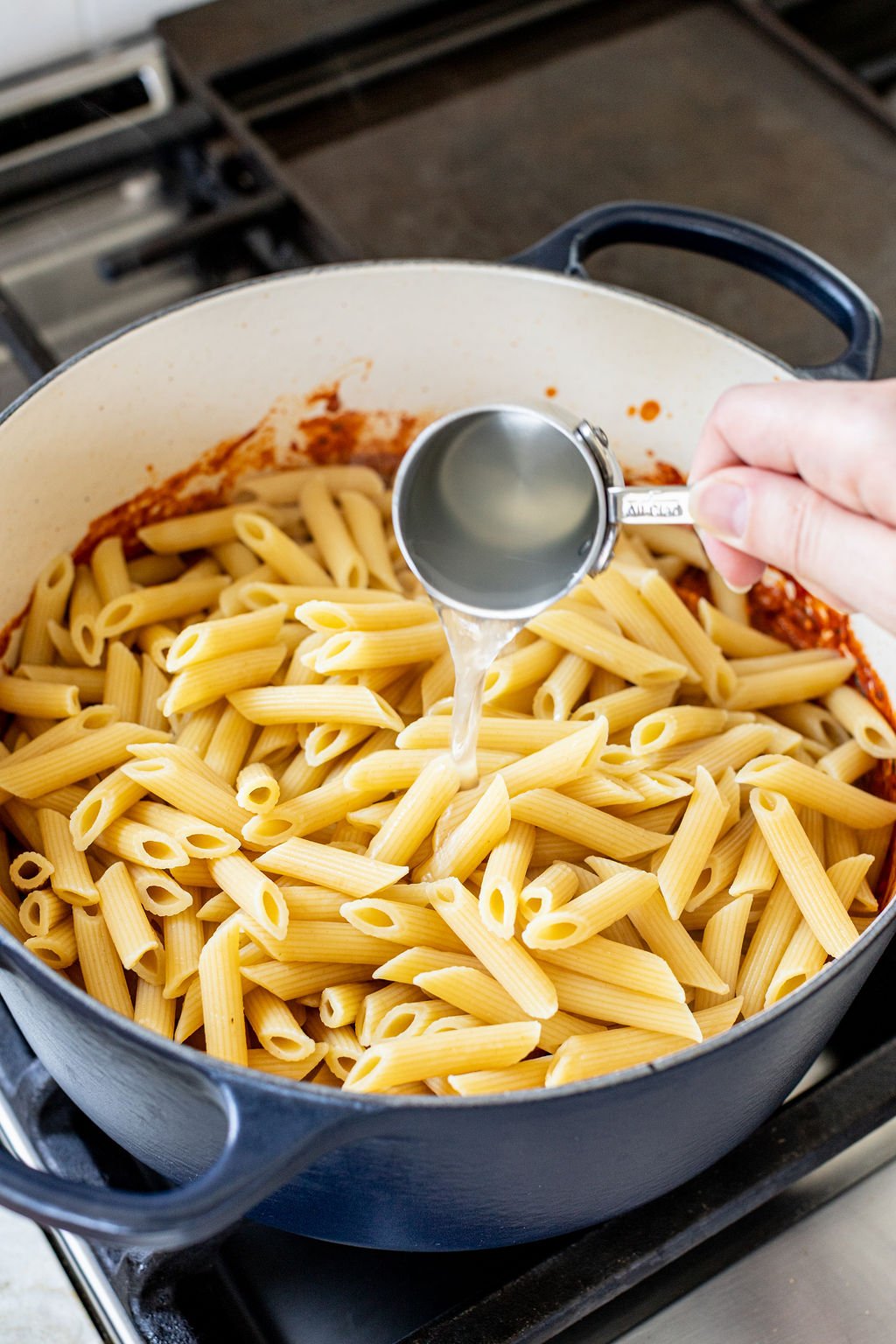 A hand pouring water onto cooked pasta in a pot with red sauce.
