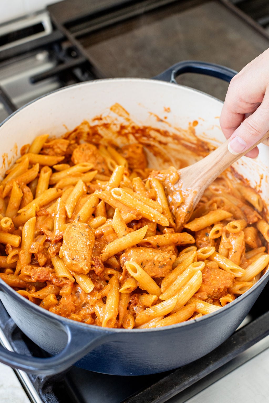 A hand stirring creamy pasta with chicken and sun dried tomatoes in a pot.